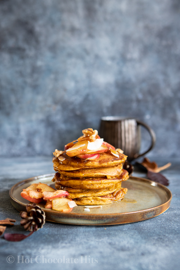 A stack of pumpkin pancakes (centre), coffee mug in the background, pine cones and fall leaves in the foreground and background.