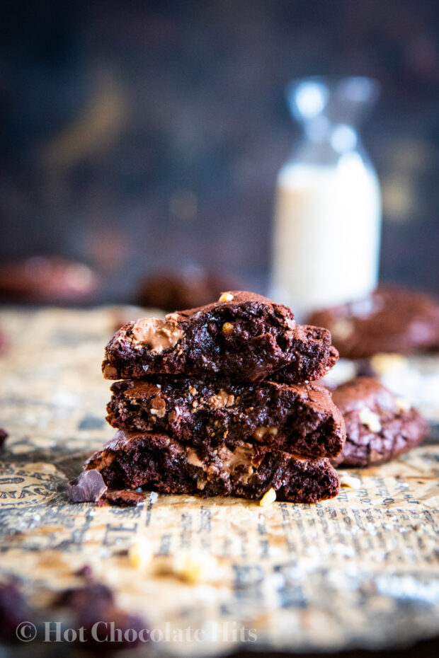 dark background, a small bottle of milk. In the foreground, three cookie halves stacked on top of each other to show the inside. Melted chocolate comes out. More cookies in the background.