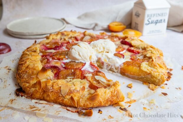A rustic plum tart (center) with scoops of vanilla ice cream. A piece has been cut out. Plates and plums pie in the background.