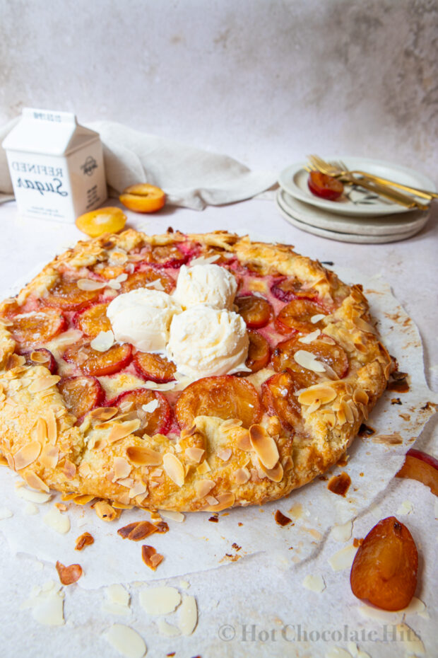 A rustic plum tart (center) with scoops of vanilla ice cream. Plates and plums pie in the background.